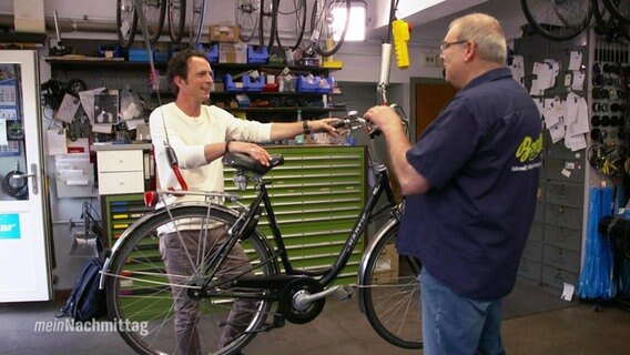Jo Hiller und ein weiterer Mann stehen in einer Fahrradwerkstatt vor einem zur Reperatur aufgehängten Fahrrad.  