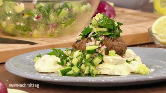 Auf einem Teller liegt eine Frikadelle auf Kartoffelsalat, dekoriert mit Gurke, Petersilie und Zwiebeln.  