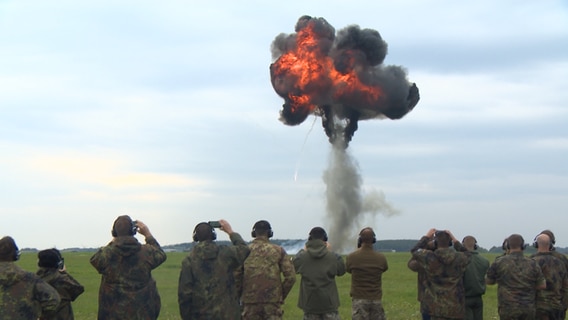 Eine Gruppe von Soldaten und Soldatinnen steht vor einer Wiese von der aus eine Rauchwolke in den Himmel steigt.  