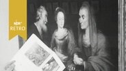 Ein Ausstellungskatalog wird vor ein Gemälde mit zwei jungen Frauen und einem bärtigen älteren Mann gehalten (1964)  
