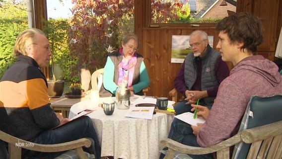 Vier Personen sitzen um einen Tisch in einem Wintergarten.  