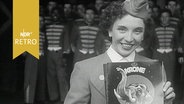 Ansagerin präsentiert Programmheft des Zirkus Krone vor Zirkusmitarbeitern in Uniformen (1956)  