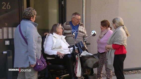 Mehrere Menschen unterhalten sich miteinander vor einer Haustür, eine ältere Dame sitzt in einem Rollstuhl.  