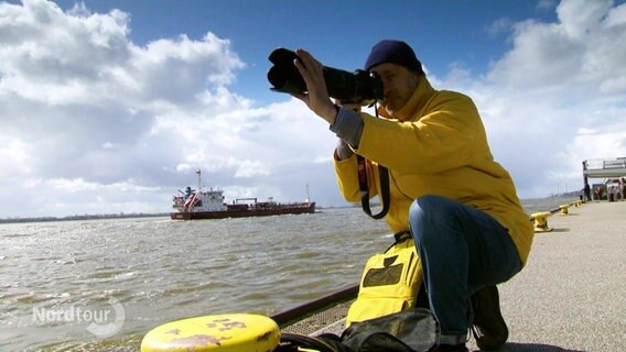 Schiffsfotograf Thomas Kunadt in gelber Öljacke und blauer Strickmütze auf einem Anleger, er schaut durch die Linse seiner Fotokamera. Im Hintergrund ein Schiff der Berufsschifffahrt.  