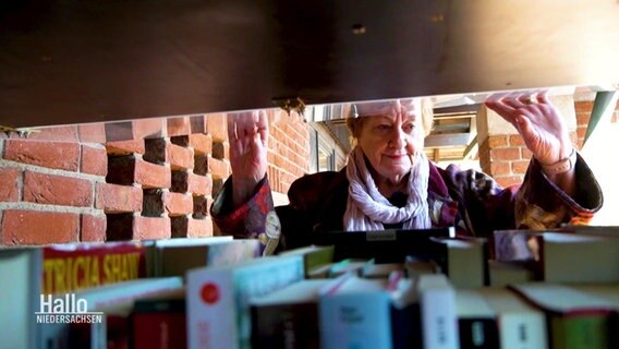 Helga Timm-Koltermann hebt eine Klappe nach oben und schaut in den Bücherschrank. Im Vordergrund stehen Bücher.  