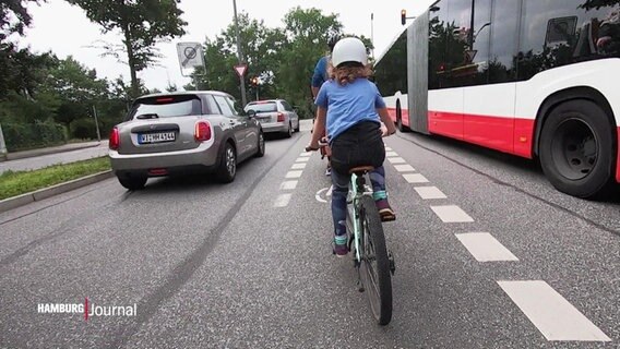 Von hinten ist ein Kind mit Helm auf einem Fahrrad zu sehen. Der Fahrradstreifen führt zwischen der Busspur und der Autospur entlang.  