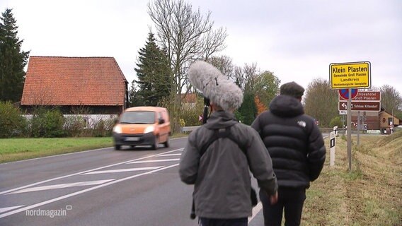 Zwei Männer gehen am Rande einer Landstraße auf ein Dorf zu, auf dem Ortsschild steht Klein Plasten.  