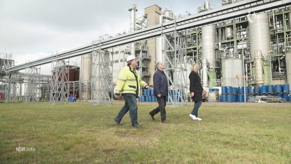Drei Personen laufen über eine Wiese vor einer Industrieanlage.  
