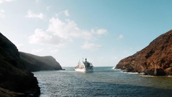 Von hinten zu sehen: EIn Versorgungsschiff fährt durch die Südsee, vorbei an felsigen Inseln. Das Wasser ist türkisblau, die Sonne scheint.  