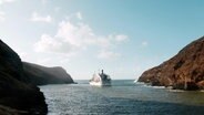 Von hinten zu sehen: EIn Versorgungsschiff fährt durch die Südsee, vorbei an felsigen Inseln. Das Wasser ist türkisblau, die Sonne scheint. © Screenshot NDR 
