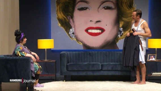 Eine Schauspielerin und ein Schauspieler stehen in ihren Rollen auf der Bühne von "Weddersehn maakt Freid". Die Bühne ist hergerichtet wie ein Wohnzimmer, an der hinteren Wand ist ein übergroßes Bild einer blonden Frau im Pop-Art-Stil.  