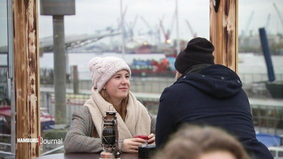 Ein Mann und eine Frau mit Mützen genießen ein heißes Getränk mit Blick auf den Hamburger Hafen.  