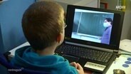 Ein Schüler schaut auf einem Laptop einem Lehrer an der Tafel zu  