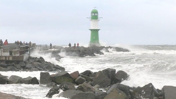 Bilder von der Nordsee während dem Sturmtief Daniel: Wellen brechen sich an der Küste und Brandung tobt um einen grün-weißen Leuchtturm. Einzelne Spaziergänger.  