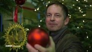 Profi-Schmücker Marcel Klöpner hält eine rote, matte Weihnachtskugel vor die Kamera. © Screenshot 