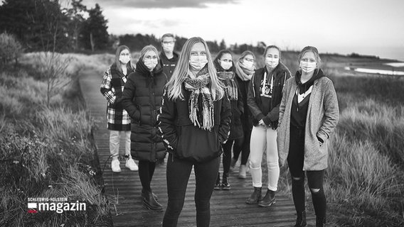 Eine Fotografie in schwarz-weiß: Mehrere Jugendliche stehen auf einem Steg zwischen kleineren Dünen. Sie alle tragen eine Maske über Mund und Nase. © Screenshot 
