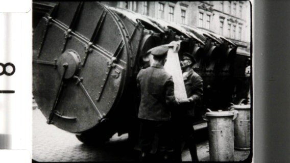 Mitarbeiter der Müllabfuhr kippen auf einem Schwarz-Weiß-Bild den Inhalt von Mülltonnen in ein Transportfahrzeug. © Screenshot 