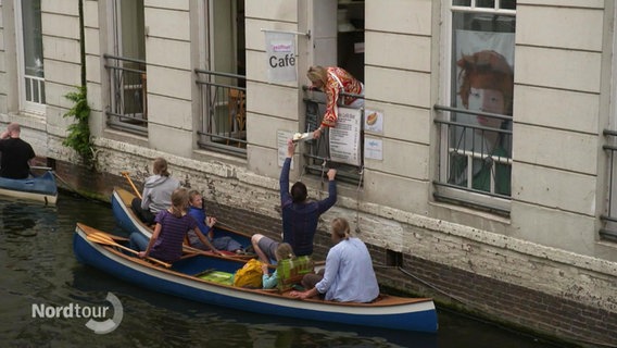 Aus einem Fenster aus einem Haus an einem Kanal, reicht eine Frau etwas an Paddler weiter. © Screenshot 