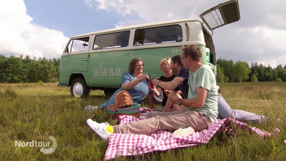 Vier Menschen machen ein Picknick in der Heide, im Hintergrund steht ein VW-Bulli. © Screenshot 