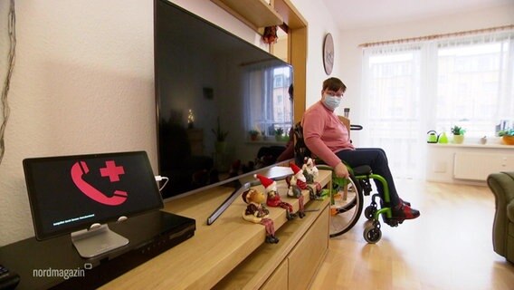 Eine junge Frau fährt mit ihrem Rollstuhl durch eine Wohnung und schaut auf ein blinkendes Display. © Screenshot 