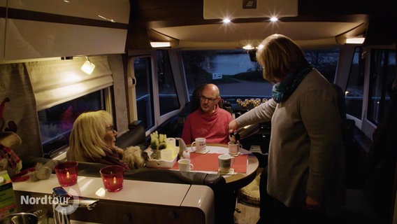 In der Sitzecke eines Wohnmobils schenkt eine Frau zwei anderen Personen ein Heißgetränk ein. © Screenshot 
