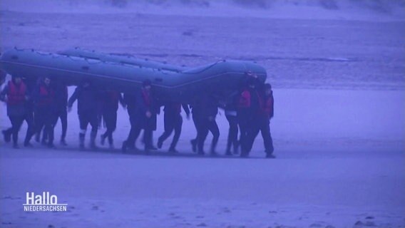 Flüchtende Menschen tragen ein Schlauchboot an einem Strand zum Meer. © Screenshot 