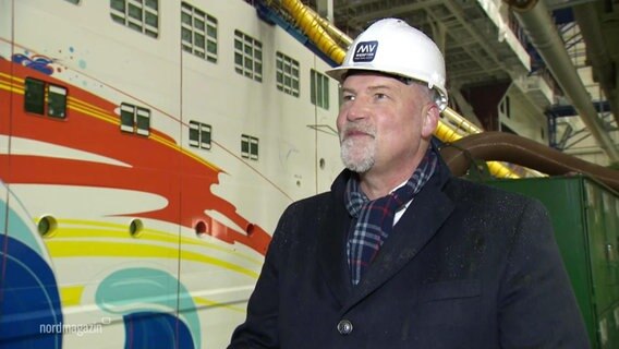 Carsten Haake, Geschäftsführer MV-Werften, steht vor einem Kreuzfahrtschiff, das in einer Halle gebaut wird. © Screenshot 