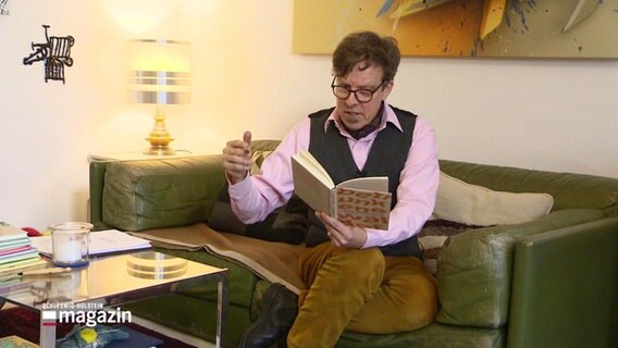 Der Dichter Arne Rautenberg sitzt auf einem Sofa und liest aus einem Buch vor. © Screenshot 