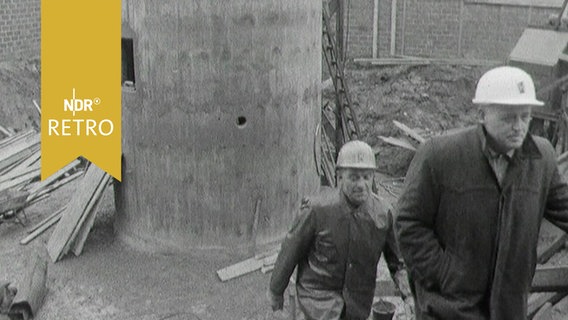 Zwei Männer mit Bauarbeiterhelmen gehen eine Bautreppe hoch, im Hintergrund der untere Teil eines Leuchtturms (1963) © Screenshot 