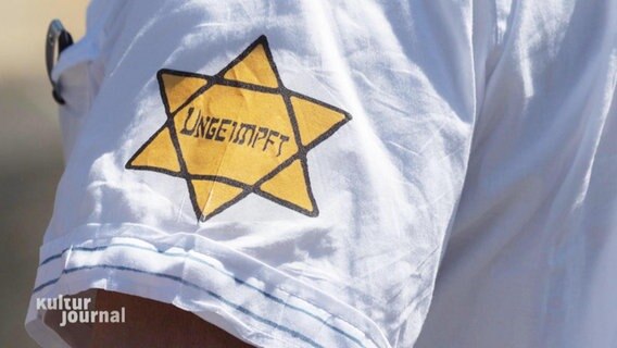 Nahaufnahme von einem weißen T-Shirt-Ärmel. Darauf befindet sich ein gelber Davidstern im Stil des Judensterns im Dritten Reich. Darin steht: "Ungeimpft". © Screenshot 