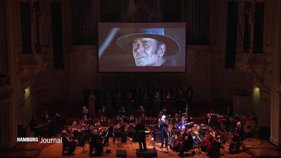 Ein Orchester spielt in der Hamburger Laeiszhalle, im Hintergrund wird eine Szene aus Zwei glorreiche Halunken projiziert. © Screenshot 