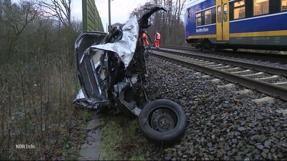 Ein zerstörtes Auto am Rande eines Gleisbettes, dahinter ein Zug. © Screenshot 