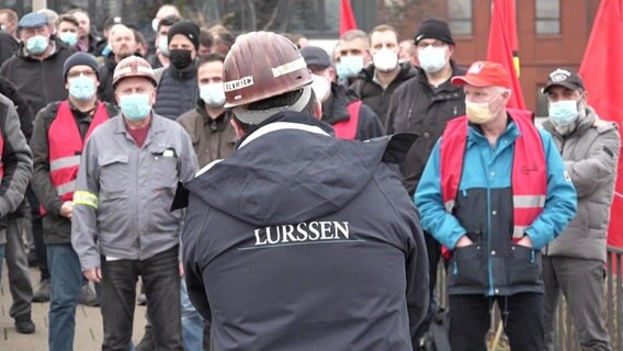 Ein Mann mit einer Jacke mit der Aufschrift "LÜRSSEN" spricht mit Werftarbeitern mit Atemschutzmasken © Screenshot 