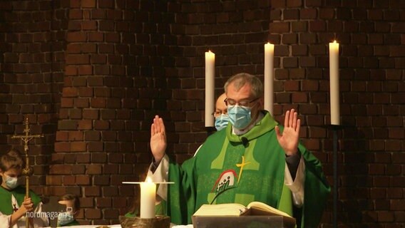 Ein Pfarrer während eines Gottesdienstes am Altar, er breitet die Hände aus und liest in der Bibel. Er trägt eine Schutzmaske. © Screenshot 