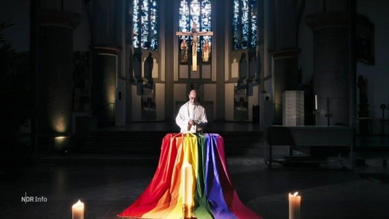 Ein Priester steht vor einer Regenbogenflagge in einer Kirche. © Screenshot 