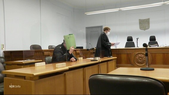 Ein Angeklagter verbirgt sein Gesicht hinter einem Aktenordner. © Screenshot 