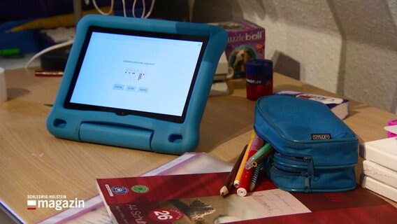 Ein Tablet steht auf einem Schülerschreibtisch, daneben eine Federtasche und Notizblöcke. © Screenshot 