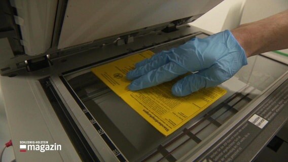 Eine Handschuh tragende Hand legt einen Impfausweis auf die Kopierfläche eines Druckers © Screenshot 