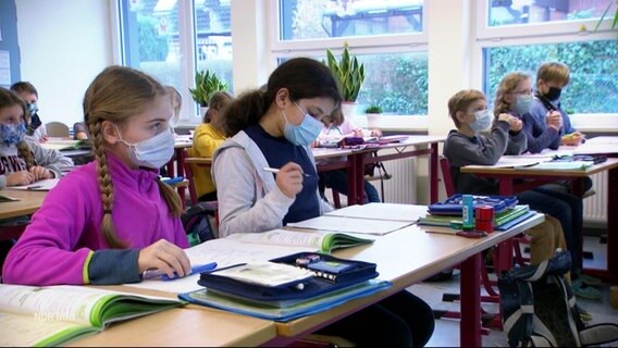 Zwei Schülerinnen sitzen in der ersten Reihe in einem Klassenzimmer. Sie tragen Corona-Masken. © Screenshot 