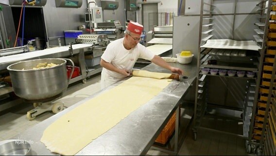 Ein Bäcker rollt einen Teig auf einer längeren Arbeitsfläche aus. © Screenshot 