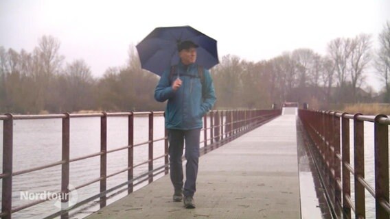 Ein Mann mit Regenschirm spaziert auf einem Steg. © Screenshot 