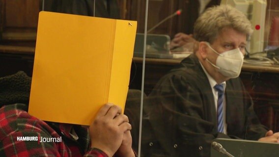 Eine Person in einem Gerichtssaal hält sich eine Mappe vors Gesicht. © Screenshot 