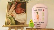 Die kindgerecht gestaltete Urne von Juna Marie. Daneben steht ein Plüsch-Wellensittich und ein Foto von ihr. © Screenshot 