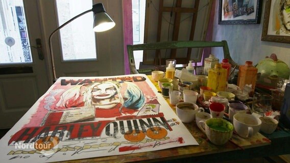 Auf einem Tisch liegt eine unvollendete Collage, daneben zahlreiche Tassen mit Farbe. © Screenshot 