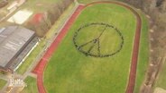 Blick aus der Vogelperspektive: Auf einem Sportplatz haben sich viele SchülerInnen zu einem reisigen Peace-Symbol formiert. © Screenshot 