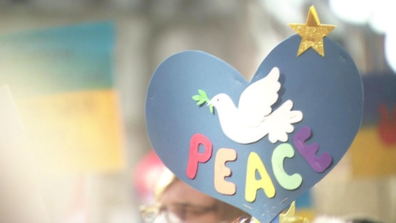 Ein gebasteltes blaues Herz. Darauf eine weiße Friedenstaube aus Papier unter der "Peace" steht. © Screenshot 