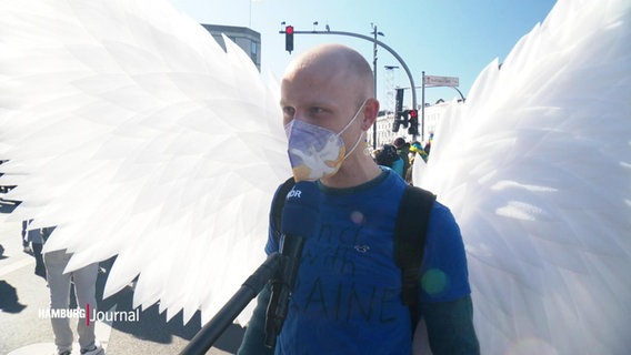 Ein Demonstrant mit weißen Flügeln im Interview. © Screenshot 