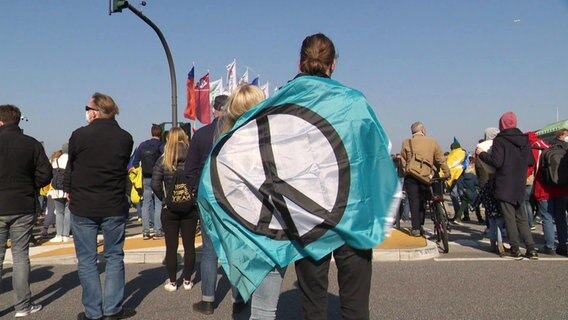 ZWei Menschen von hinten, umhüllt mit einem Tuch, auf dem ein Peace-Zeichen abgebildet ist © Screenshot 
