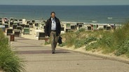 Ein Mann mit Aktentasche läuft an einer Strandpromenade. © Screenshot 