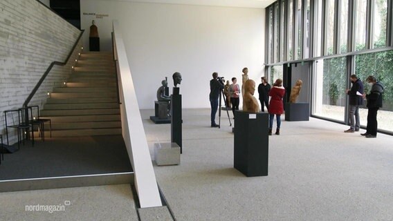 In einem weißgestrichenen, lichtgefluteten größeren Raum mit breiter Fensterfront stehen mehrere Büsten und Skulpturen auf schwarzen Podesten sowie einige Besucher der Ausstellung. © Screenshot 